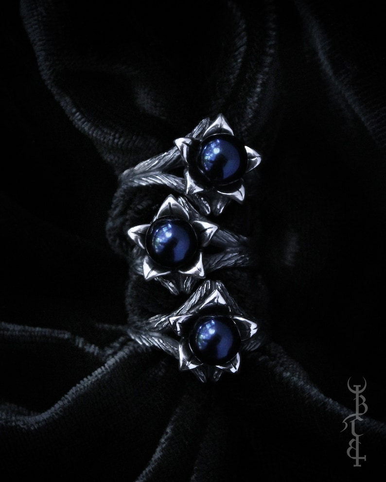 Op de afbeelding: Drie zilveren ringen met zwarte parel accenten. Elke ring heeft een gestileerd bloemendesign met een enkele parel in het midden.
