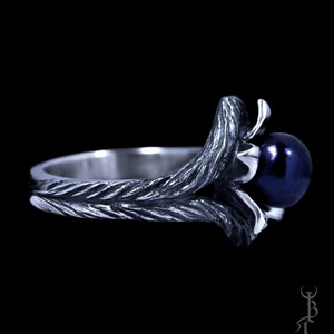 Op de afbeelding: Een zilveren ring met een donkerblauwe parel gezet in een gedraaid takontwerp.