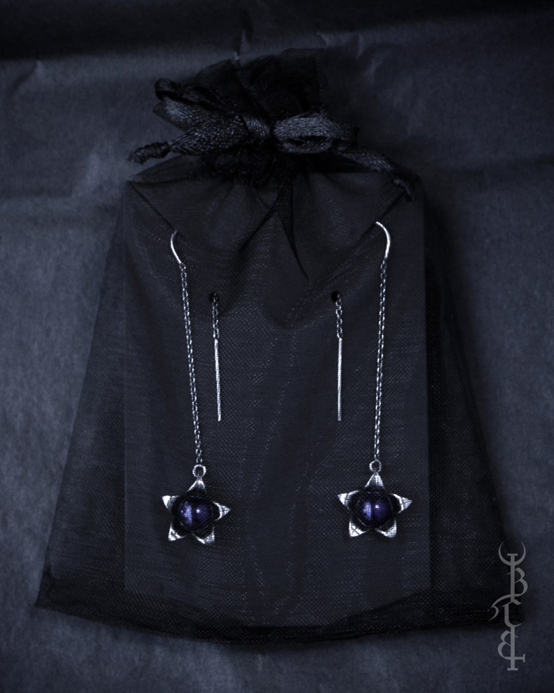 May include: A pair of silver star-shaped earrings with dark purple beads hanging from a black velvet bag.