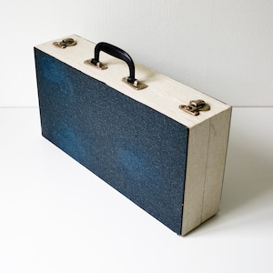May include: A rectangular carrying case featuring a dark blue textured fabric exterior on the front and back panels, contrasted by light beige side panels. The case includes a black plastic handle and two metallic latches on the top edge.