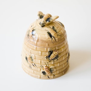 May include: A tan ceramic honey pot shaped like a traditional skep beehive with a removable lid. The exterior features a raised, textured basket-weave pattern and several sculpted, painted bees with black and brown details. A single bee sits on top.