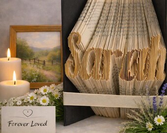 May include: A folded book art sculpture with the name Sarah spelled out through precisely creased pages. The book features a black cover and a cream-colored ribbon band. A small white card in front displays a heart icon and the text Forever Loved.