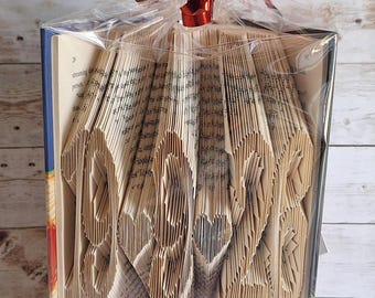 May include: A folded book art sculpture featuring the numbers 1926 with two heart shapes between them. The pages are intricately creased to form the design. The book is wrapped in clear plastic with a shiny red ribbon bow on top and a red band.