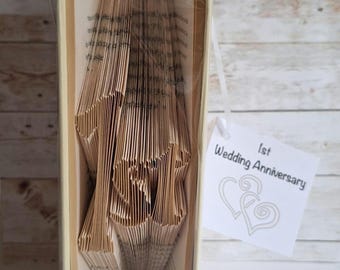 May include: A book with pages folded to form the text 1st, wrapped in clear plastic with a white ribbon and a white tag that reads 1st Wedding Anniversary with two heart illustrations. The book is held together by a white band at the base.