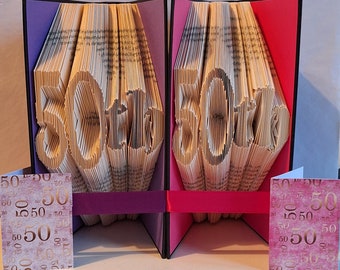 May include: Two book folding sculptures, each shaped like the number 50, are displayed on a table. The sculptures are made from pages of a book and are folded into a three-dimensional shape. The sculptures are painted in shades of pink and purple.
