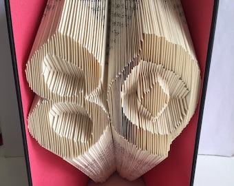 May include: A book sculpture featuring folded pages that form the number 80. The book is set against a bright pink interior cover and a black outer spine, with a horizontal pink ribbon wrapped around the base of the open book structure.