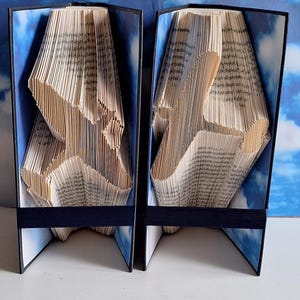May include: Two book sculptures of birds in flight, each made from a book with pages folded into a three-dimensional shape. The books are bound in black and have a blue sky background. The sculptures are standing on a white surface.