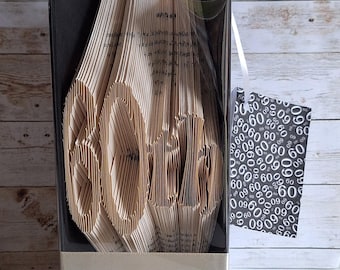 May include: A book folded into the shape of the number "60" as a gift for a 60th birthday. The book is wrapped in clear plastic and tied with a white ribbon. A small gift tag with a black and white pattern of the number "60" is attached to the gift.