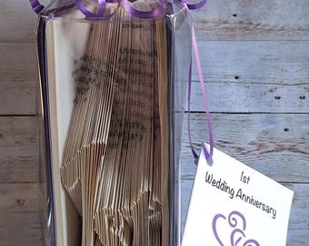 May include: A folded book art sculpture forming the word 1st, wrapped in clear plastic with a purple ribbon bow on top. A white square tag attached with a purple string reads 1st Wedding Anniversary with two purple heart illustrations.