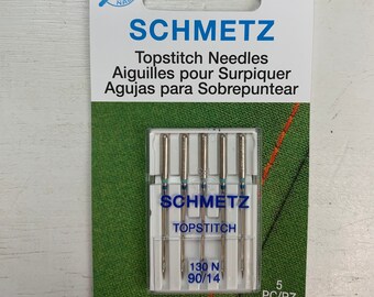 May include: A pack of 5 topstitch needles for sewing machines. The needles are silver with blue tips and are labeled "SCHMETZ TOPSTITCH 130 N 90/14".