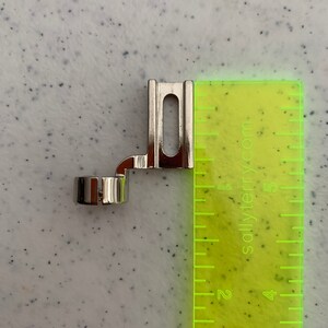 May include: A silver sewing machine foot attachment for creating rolled hems. The foot is about 1.5 inches long and has a small, round opening for the fabric.