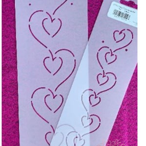 May include: Two white plastic quilting stencils with a design of a row of hearts. The stencils are on a pink fabric background.