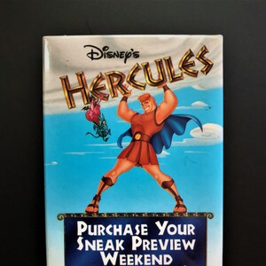 May include: A vintage Disney's Hercules movie promotional magnet. The magnet features a cartoon image of Hercules lifting a large rock with the words "Disney's Hercules" above him. The text "Purchase Your Sneak Preview Weekend Tickets Here!" is printed on a blue banner below Hercules.