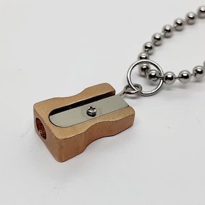 May include: A gold-coloured metal pencil sharpener with a silver blade is attached to a silver chain. The sharpener is a small, portable item that would be perfect for a keychain.