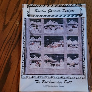 May include: A package for a quilt pattern titled "The Breckenridge Quilt" by Shirley Gardner Designs. The quilt design features snowy mountain scenes with houses, trees, and horses. The package has the text "Shirley Gardner Designs" at the top.