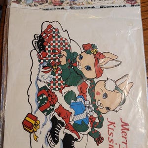 May include: A Cache Junction iron-on transfer package featuring two cartoon rabbits ice skating. The rabbits wear winter clothing and hold wrapped gifts. The text reads Cache Junction Iron-on Transfers by Seitec and Merry Kissmas.