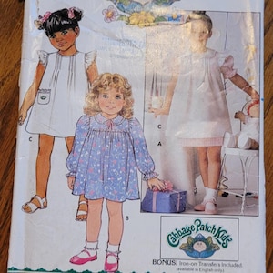 May include: A vintage sewing pattern for Cabbage Patch Kids clothing. The pattern includes instructions for making two dresses, one white and one blue with a floral print. The pattern is from Butterick and has the number 5595 on it.
