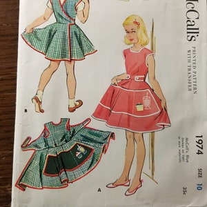 May include: A vintage McCall's sewing pattern for a girl's wrap-around cobbler apron. The pattern is printed on white paper with a pink and green checkered design. The pattern includes a transfer sheet and is size 10. The pattern is for a girl's wrap-around cobbler apron. The pattern is printed on white paper with a pink and green checkered design. The pattern includes a transfer sheet and is size 10.