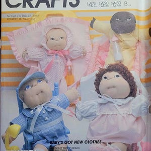 May include: A sewing pattern for making clothes for 12-inch dolls. The pattern includes instructions for making a pink dress, a blue dress, and a yellow outfit. The pattern is called "Baby's Got New Clothes..." and "Bebe a de Nouveaux Habits..." McCall's Crafts 9581.