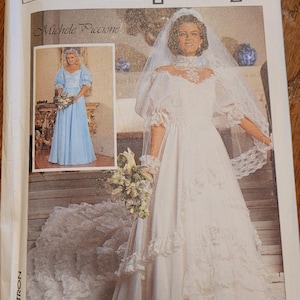 May include: Simplicity sewing pattern featuring a white wedding dress with a high neckline, lace details, and a tiered skirt. The pattern includes a smaller image of a blue dress. The pattern is labeled with the number 12 and the name "Michele Piccione".