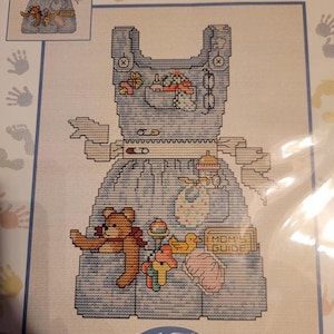 May include: A cross-stitch pattern featuring a light blue apron decorated with various baby-themed items including a teddy bear, a yellow rubber duck, a baby bottle, a bib, and a book titled MOM'S GUIDE, all rendered in colorful embroidery thread.