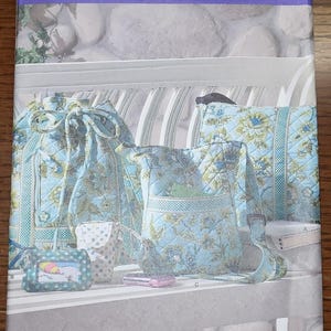 May include: Simplicity pattern 2911 featuring a collection of quilted bags and accessories in a floral pattern. The set includes a tote bag, drawstring bag, pillow, and small pouches. The color scheme is primarily light blue with green and white floral accents.