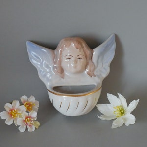 May include: A ceramic wall planter shaped like an angel with wings, a head, and a small planter in the front. The angel is painted in shades of blue, white, and brown. The planter has a gold trim around the top.