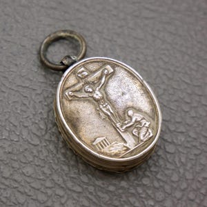 Pode incluir: Pingente oval prateado com um relevo detalhado da crucificação de Jesus Cristo. O pingente tem um pequeno anel na parte superior e um design articulado. O relevo retrata Jesus na cruz, com figuras na base.