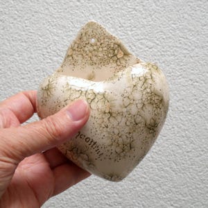 Pode incluir: Um bolso de parede de cerâmica em forma de coração, branco sujo, com um padrão texturizado e salpicado em tons de verde e castanho. O bolso tem um pequeno orifício para pendurar e a palavra "jaastrich" está escrita na lateral.