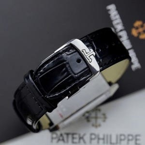 May include: A black leather watch strap with a glossy alligator texture and a polished silver-tone metal buckle. The buckle features a distinct engraved logo consisting of two interlocking J-shaped symbols. The strap is secured with a matching loop.
