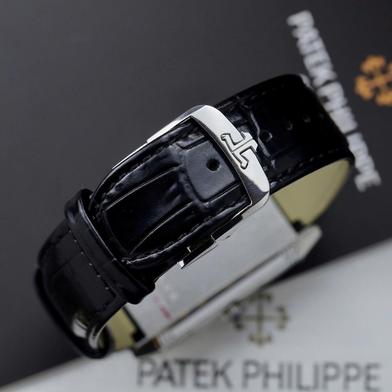 May include: A black leather watch strap with a glossy alligator texture and a polished silver-tone metal buckle. The buckle features a distinct engraved logo consisting of two interlocking J-shaped symbols. The strap is secured with a matching loop.