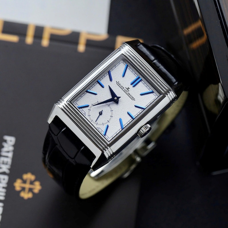 May include: A rectangular silver-tone wristwatch with a white textured dial, blue hour markers, and blue hands. The watch features a small seconds sub-dial and the text Jaeger-LeCoultre on the face. It has a black leather strap with a glossy finish.