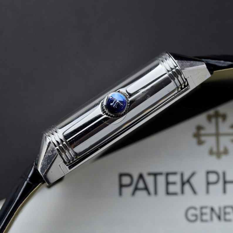 May include: A rectangular silver-toned watch case with a polished finish and ribbed edges. A small, circular blue crown with a silver logo is set into the side. The watch is attached to a black leather strap and positioned near a white card with text.