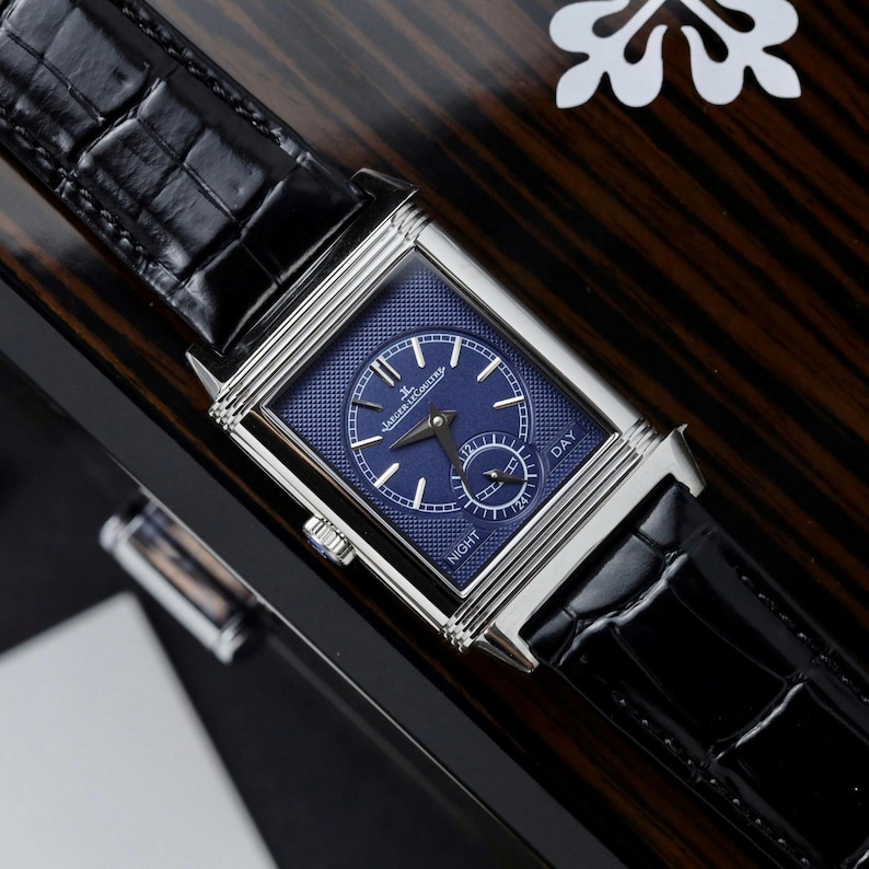 May include: A rectangular silver-tone watch with a textured blue dial, silver hands, and indices. The dial features a sub-dial with the text DAY and NIGHT, and the brand name Jaeger-LeCoultre. It has a black alligator-patterned leather strap.