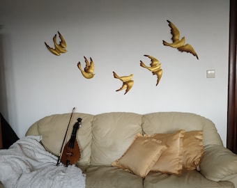 Op de afbeelding: Een lichtbeige bank met drie kussens, twee in een lichtbeige kleur en een in een donkerdere beige kleur. Een houten viool ligt op de bank. De muur achter de bank is wit met vijf gouden metalen vogelsculpturen.