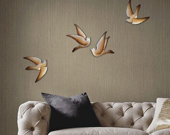 May include: Set of four metal wall decor birds in a light brown and white color scheme. The birds are in flight and are arranged in a staggered pattern on a beige wall.