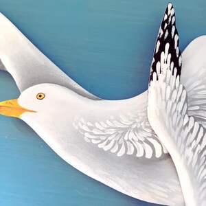 Close-up of two hand-painted white wooden seagulls with black wing tips and orange beaks, blue background - coastal wall decor