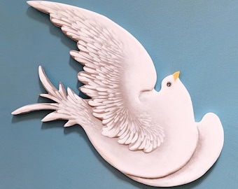 May include: A white dove sculpture with its wings spread out, flying in a circular motion. The dove is painted in a realistic style with subtle gray shading on the wings. The dove is mounted on a light blue background.