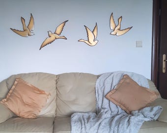 Op de afbeelding: Vier gouden houten wanddecoraties in de vorm van vogels in een vluchtformatie op een witte muur boven een beige bank met een witte deken en twee bruine kussens.