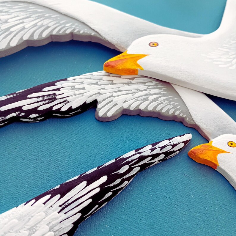 Close-up of two hand-painted white wooden seagulls with black wing tips and orange beaks, blue background - coastal nautical wall decor