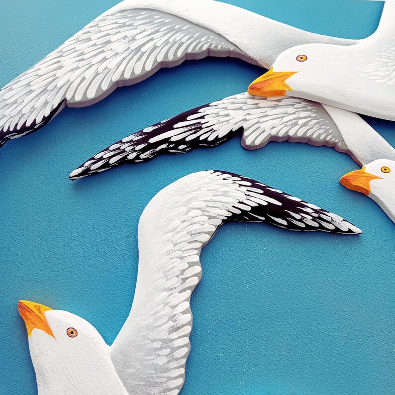 Close-up of three hand-painted white wooden seagulls with black wing tips and orange beaks, blue background.