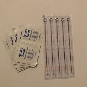 Puede incluir: Un conjunto de agujas de tatuaje en un paquete de plástico transparente, junto con varias toallitas con alcohol envueltas individualmente. Las toallitas son blancas con texto azul, y las agujas son finas, plateadas y tienen un lazo en la parte superior.