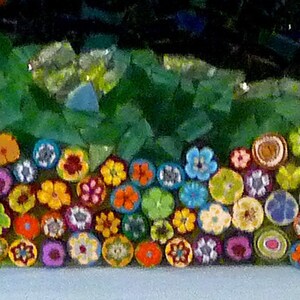 May include: A close-up of a mosaic art piece featuring a row of colorful, crocheted flowers. The flowers are arranged in a semi-circle and are made of various colors, including red, orange, yellow, green, blue, and purple.