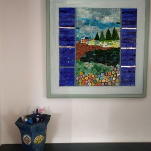 May include: A framed mosaic artwork depicting a landscape scene with a blue sky, green trees, a brown river, and colorful flowers. The mosaic is made of small, colorful tiles.