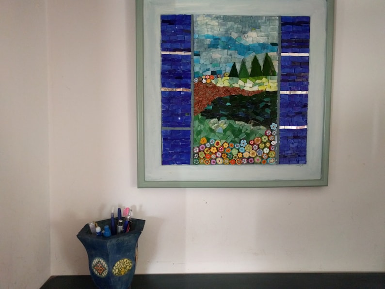 May include: A framed mosaic artwork depicting a landscape scene with a blue sky, green trees, a brown river, and colorful flowers. The mosaic is made of small, colorful tiles.