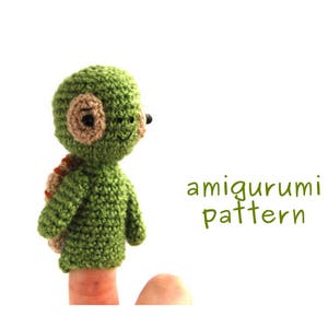 May include: Green crocheted finger puppet turtle with brown eyes and a smile. The puppet is on a finger and the text "amigurumi pattern" is below it.