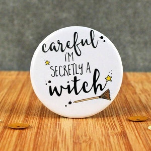 May include: A white badge with the words "Careful I'm Secretly a Witch" in black script. The badge has a yellow star and a broom illustration. It rests on a wooden surface with scattered confetti.