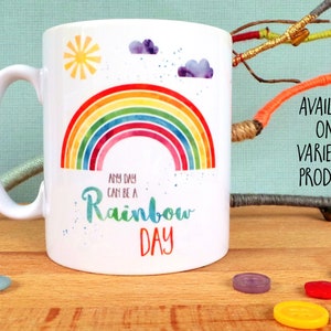 Puede incluir: Taza de cer&aacute;mica blanca con un dise&ntilde;o de arco&iacute;ris y el texto "Any day can be a Rainbow Day". La taza est&aacute; sobre una superficie de madera con botones coloridos esparcidos a su alrededor.