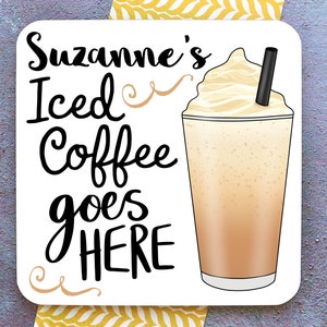 Op de afbeelding: Witte vierkante onderzetter met zwarte tekst die "Suzanne's Iced Coffee goes HERE" luidt, met een swirl-ontwerp aan beide kanten van de tekst. Rechts van de tekst staat een cartoonafbeelding van een glas met ijskoffie, slagroom en een rietje.