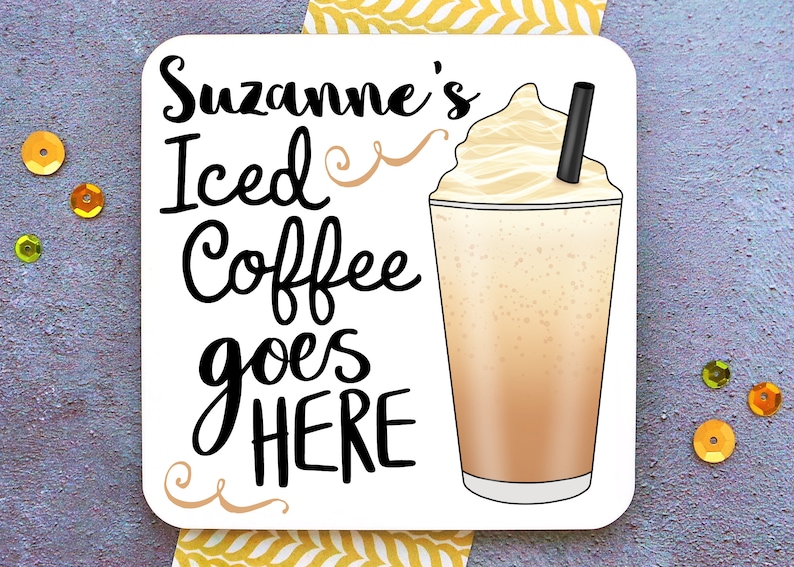 Op de afbeelding: Witte vierkante onderzetter met zwarte tekst die "Suzanne's Iced Coffee goes HERE" luidt, met een swirl-ontwerp aan beide kanten van de tekst. Rechts van de tekst staat een cartoonafbeelding van een glas met ijskoffie, slagroom en een rietje.
